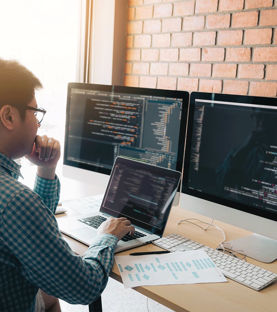 Profissional diante de um notebook e dois monitores que exibem interfaces técnicas em uma estação de trabalho.